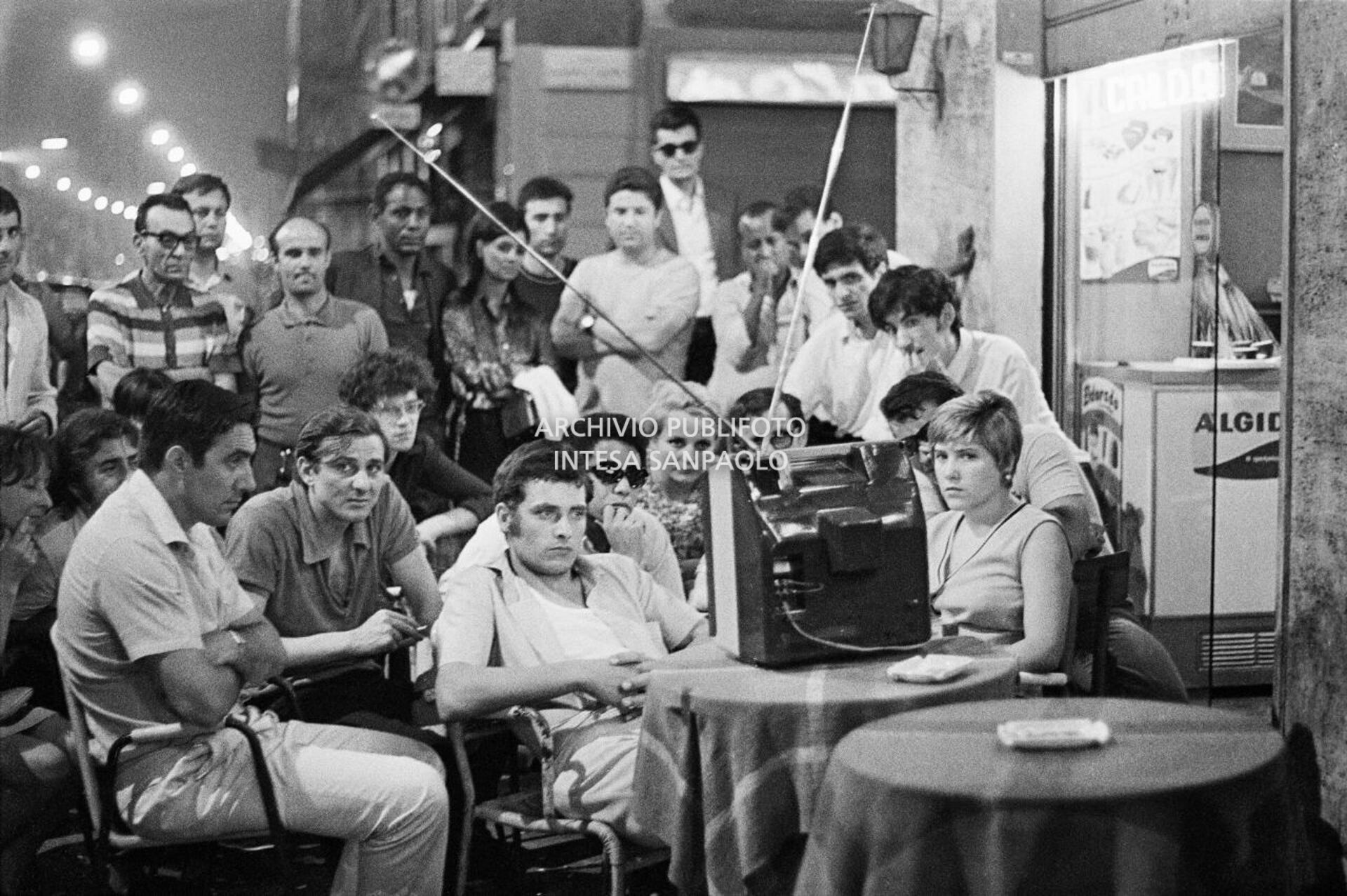 Un gruppo di persone, seduto all'esterno di un bar, segue alla televisione la missione dell'Apollo 11
