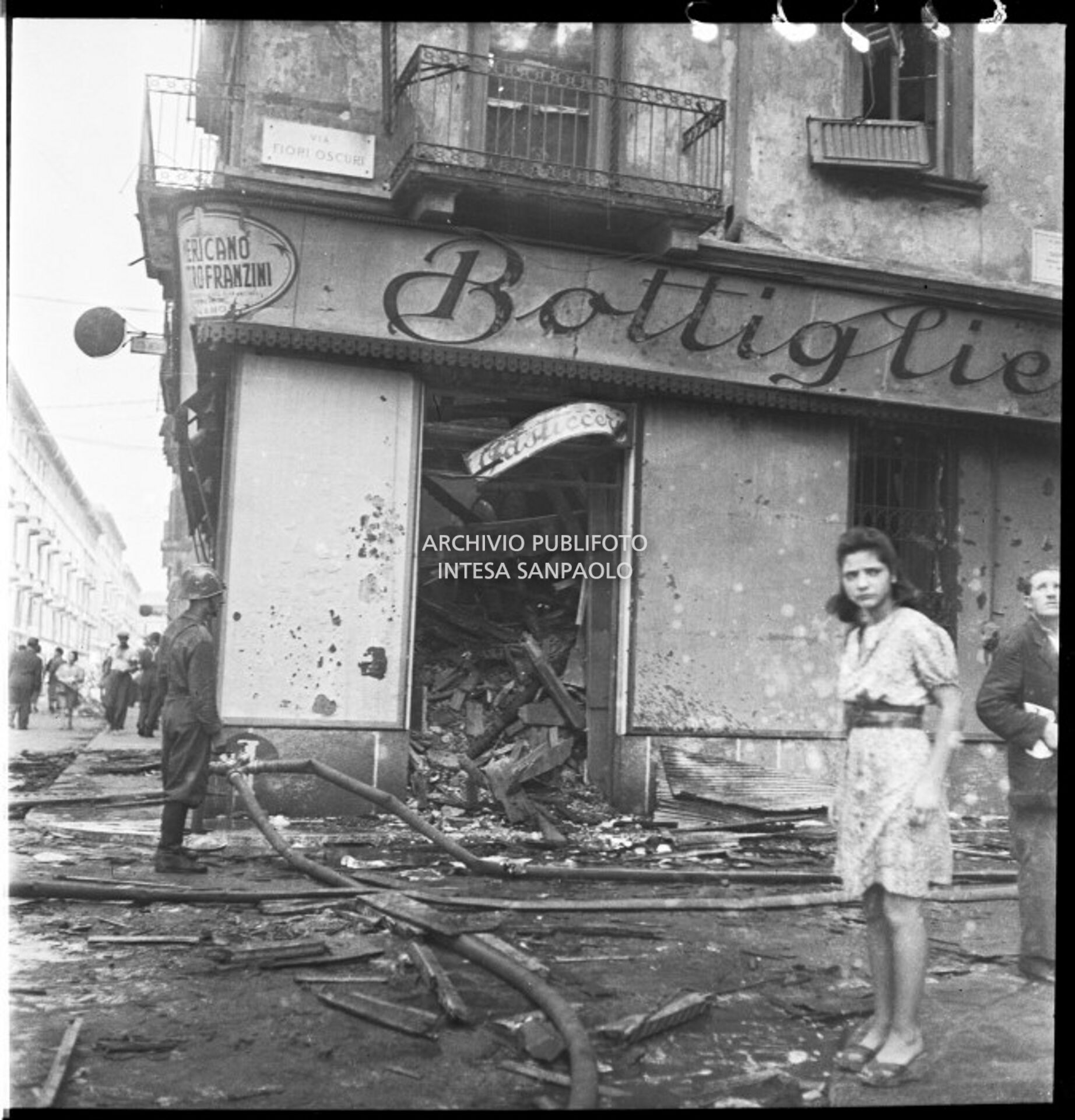 Una ragazza davanti alla bottiglieria di Luigi Piola, in via Fiori Oscuri a Milano, sventrata dai bombardamenti; a sinistra un vigile del fuoco ancora impegnato nelle operazioni di spegnimento degli incendi provocati dagli attacchi aerei