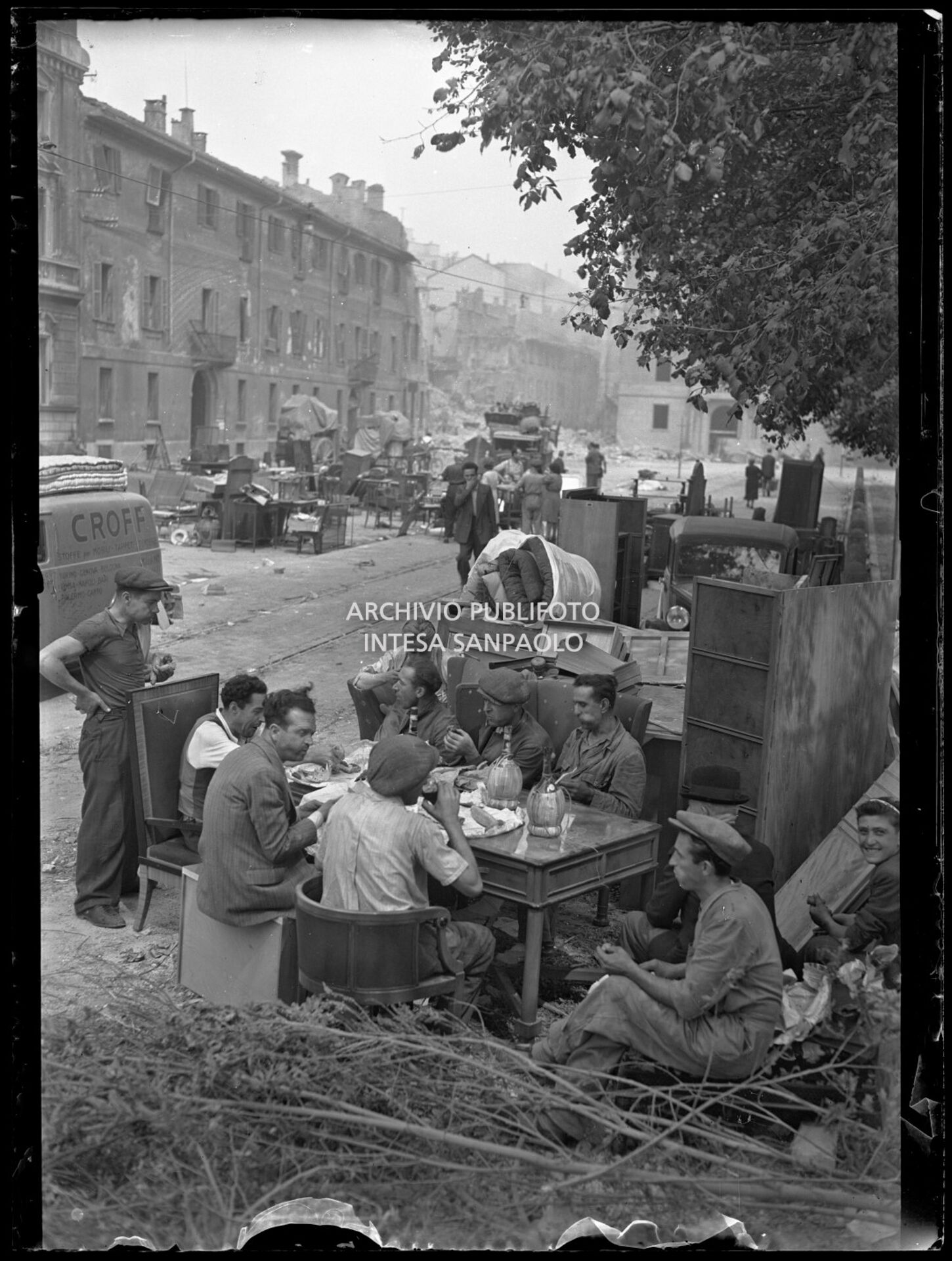 Un gruppo di uomini pranza in piazza sant'Ambrogio, a Milano, su un tavolo apparecchiato alla bell'e meglio, tra masserizie recuperate dalle case bombardate