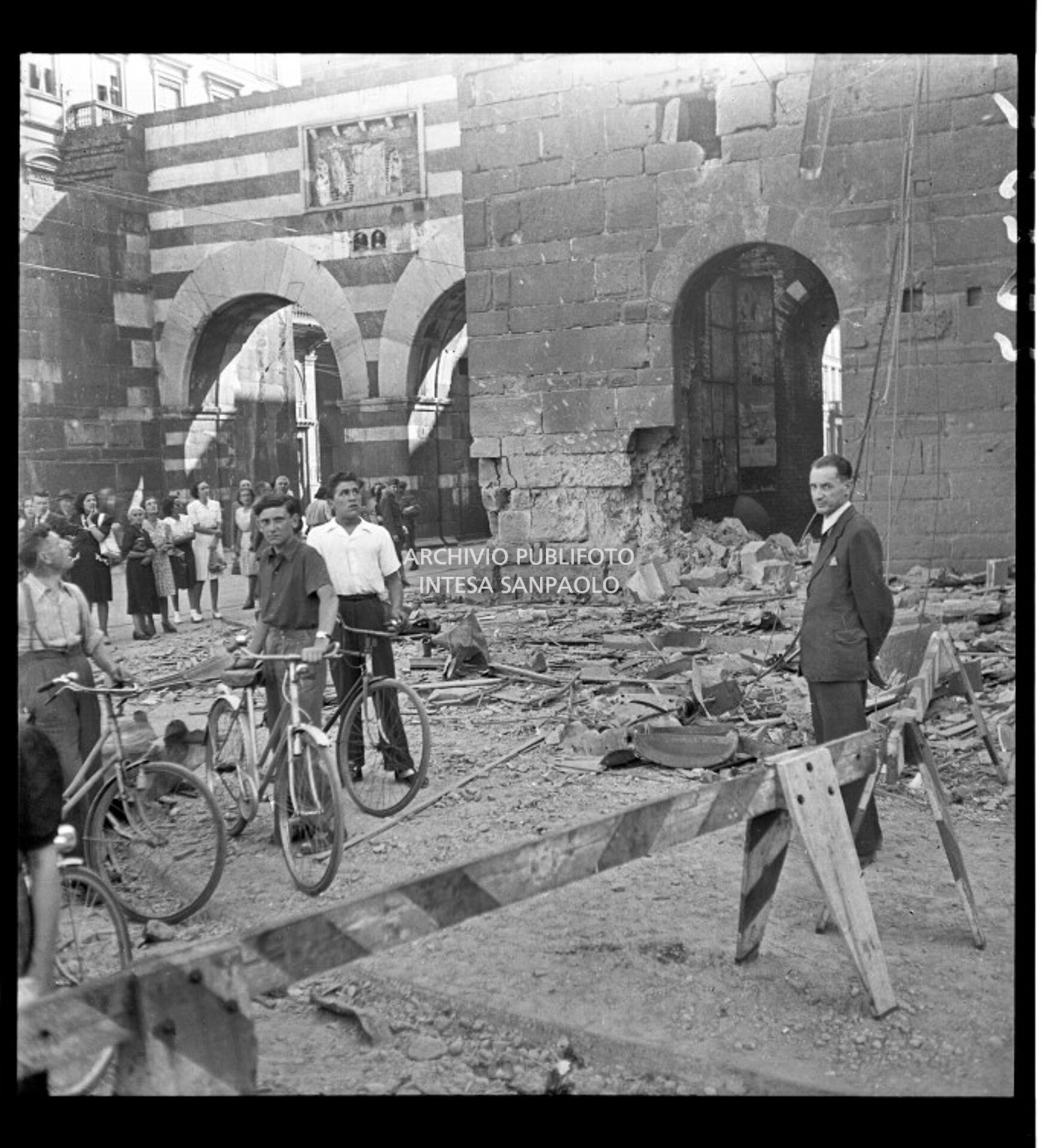 Passanti, a piedi e con la bicicletta, osservano i danni provocati dai bombardamenti agli archi di Porta Nuova medievale e a via Alessandro Manzoni a Milano