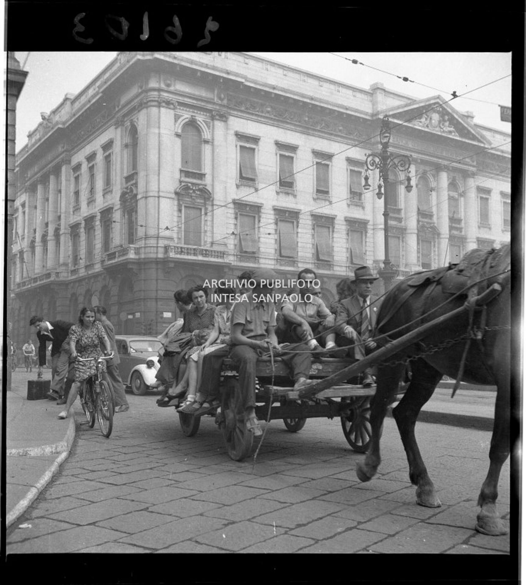 Un carro carico di persone, trainato da un cavallo, in piazza della Scala a Milano. Sullo sfondo il Palazzo della Banca Commerciale Italiana