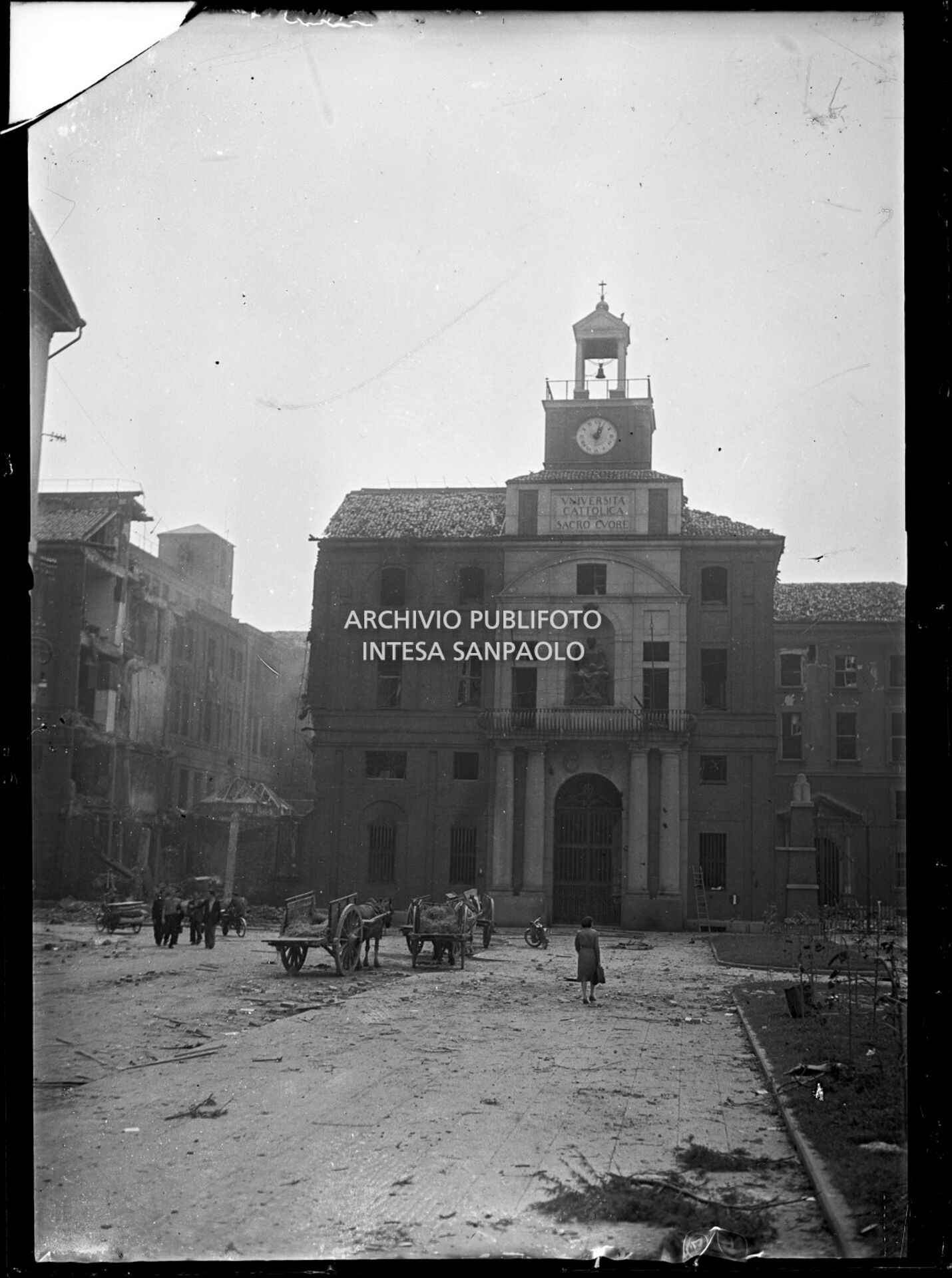 Veduta dell'edificio d'ingresso dell'Università Cattolica del Sacro Cuore, ripreso da piazza Sant'Ambrogio, a Milano, danneggiato dai bombardamenti. Due carri, con cavalli e paglia, sostano nella piazza