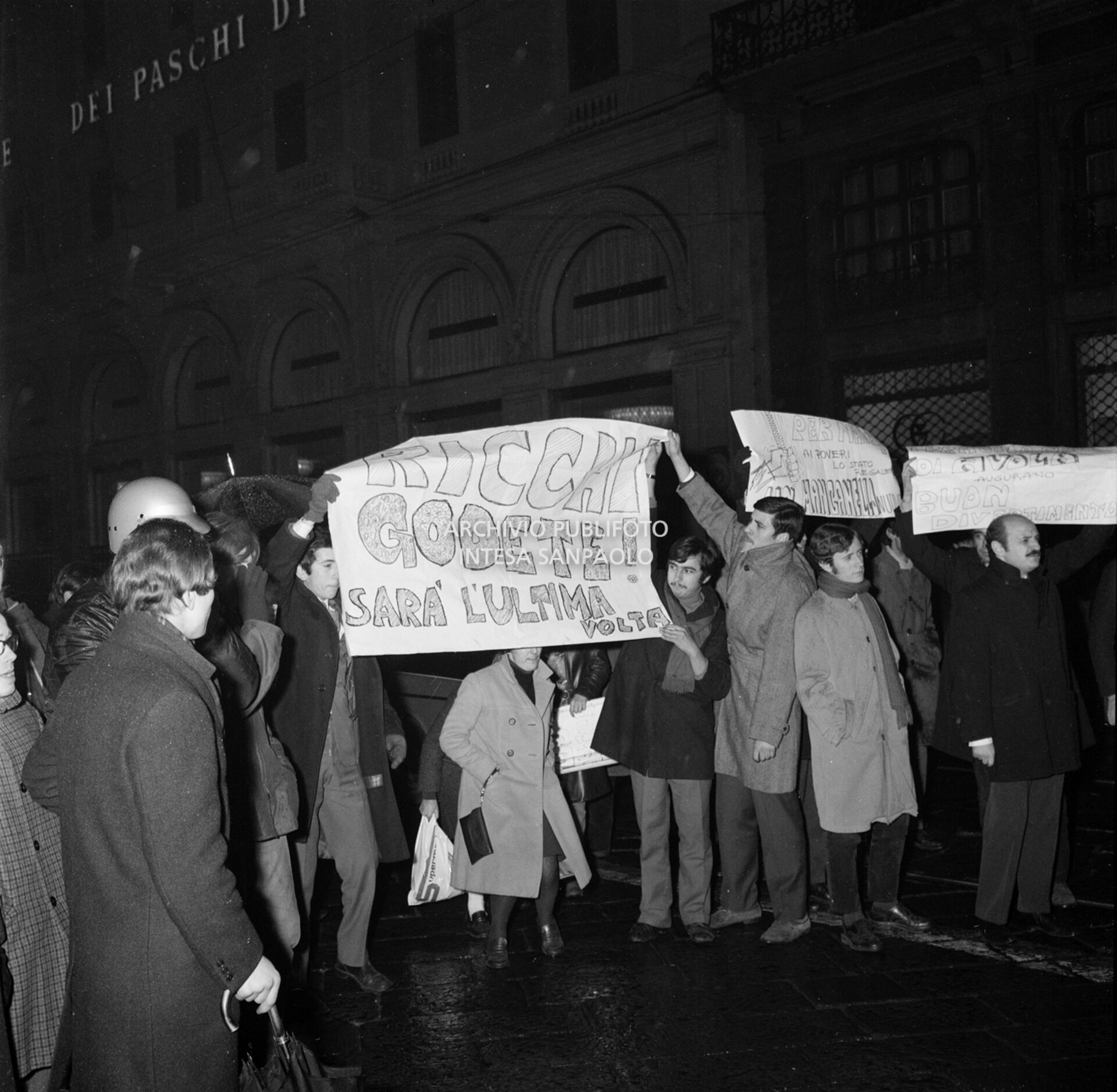 Contestatori del Movimento studentesco mostrano uno striscione con la scritta: "Ricchi godete! Sarà l'ultima volta", fuori dal Teatro alla Scala la sera dell'inaugurazione della stagione lirica 1968-1969, nel corso della quale lanciarono uova e vernice contro il pubblico in entrata