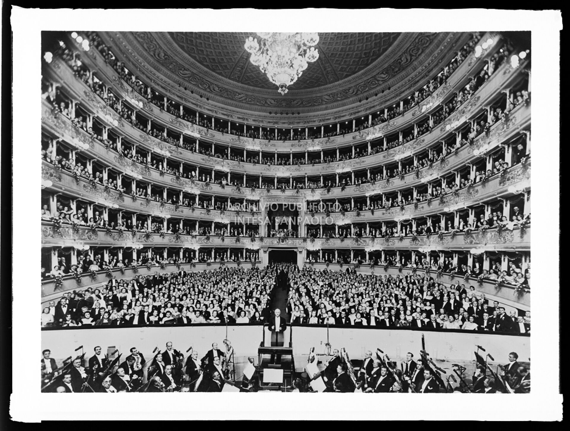 Ripresa dal palcoscenico della sala del Teatro alla Scala gremita di pubblico: sul podio il direttore Victor de Sabata, circondato dall'orchestra, la sera dell'inaugurazione della stagione lirica 1949-1950, con l'opera "La Bohème" di Giacomo Puccini, con la regia di Giovacchino Forzano