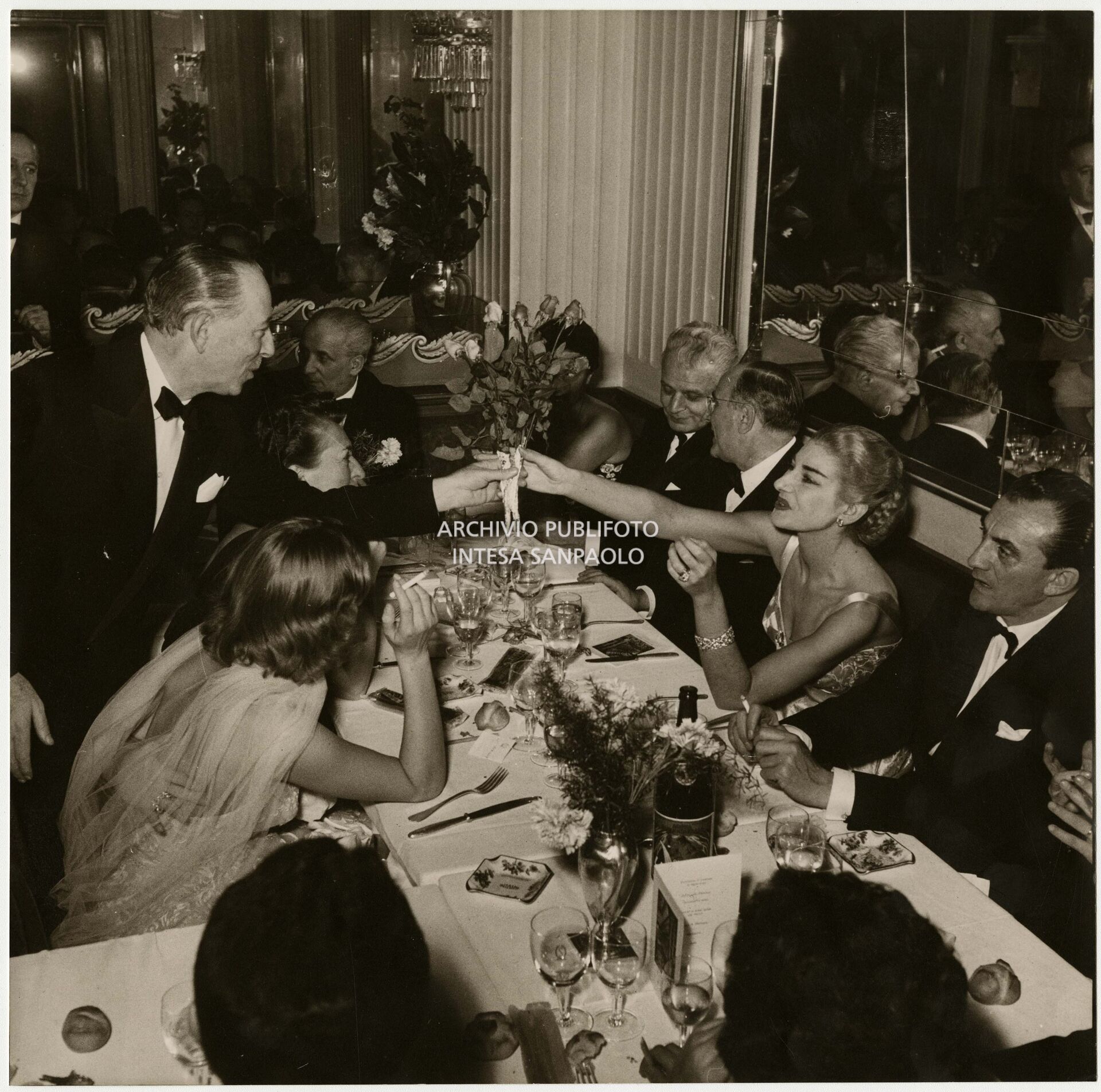 Maria Callas receives a bouquet of flowers from Valentino Bompiani, at the Savini restaurant, on the evening of the premiere of La Vestale by Gaspare Spontini. To her left, we see the director Luchino Visconti, while her husband Giovanni Battista Meneghini is at the head of the table with the superintendent of La Scala, Antonio Ghiringhelli, seated to his left.