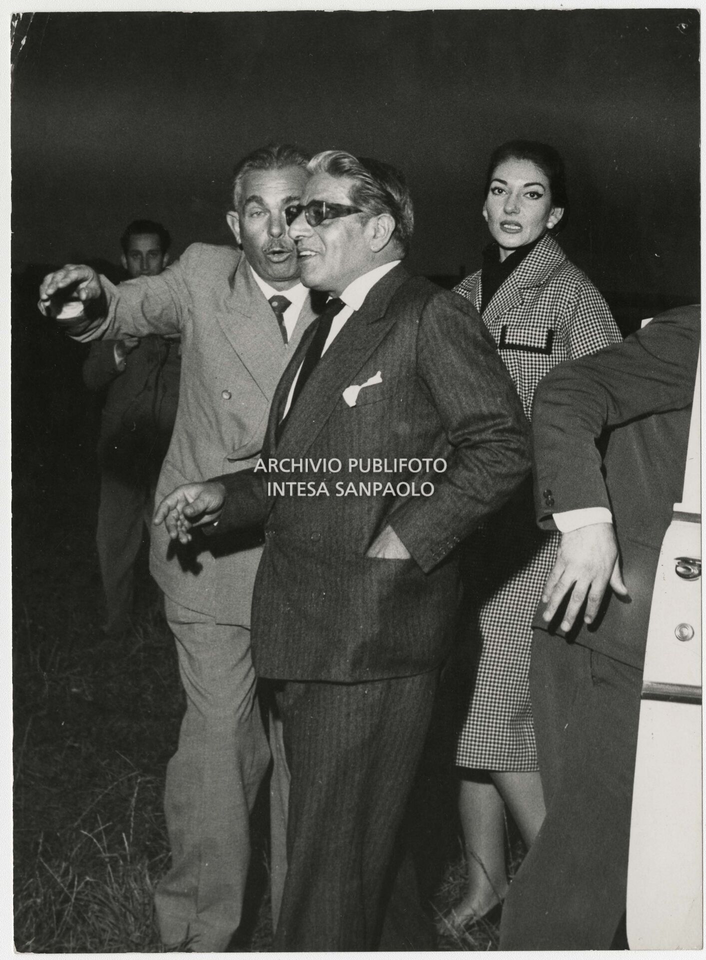 Original caption by Publifoto Agency on the back of the print:
			"One of the shots of the "Callas-Onassis affair". The public escapes of the couple that was on everyone's lips began on Saturday 29 August at the "Sea Club" in Monte Carlo. But the first photograph was taken on Thursday 3 September 1959 in Milan. Onassis arrived that afternoon from Venice in his personal plane. The two entered a Milanese night-club [the Montemerlo (Ed.)] at 10 p.m. They dined, danced, drank. They left five hours later and finally smiled at the photographer Nicola Giordano of Publifoto who was waiting for them. A short trip from the night club to the hotel, where the shipowner was staying, for a last "drink". The next day, Friday 4 September, the photographer was still waiting outside the hotel: and at 3.45 p.m. the couple came out and got into the car. Also on Friday. After a stop at the singer's house, here we are at Bresso airport. Maria Meneghini Callas says goodbye to the shipowner who is leaving for Venice in his seaplane. The few people present notice that the lady "is very emotional"."