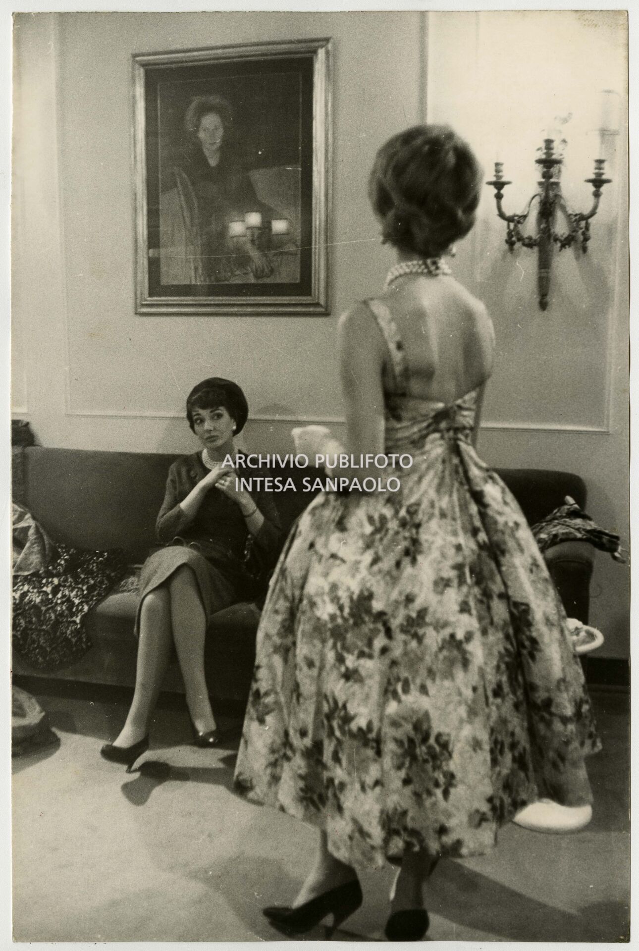 Maria Callas nell'atelier Biki ammira gli abiti che le mostra un'indossatrice; sulla parete un quadro in cui è ritratta la stilista Elvira Leonardi Bouyeure, detta Biki