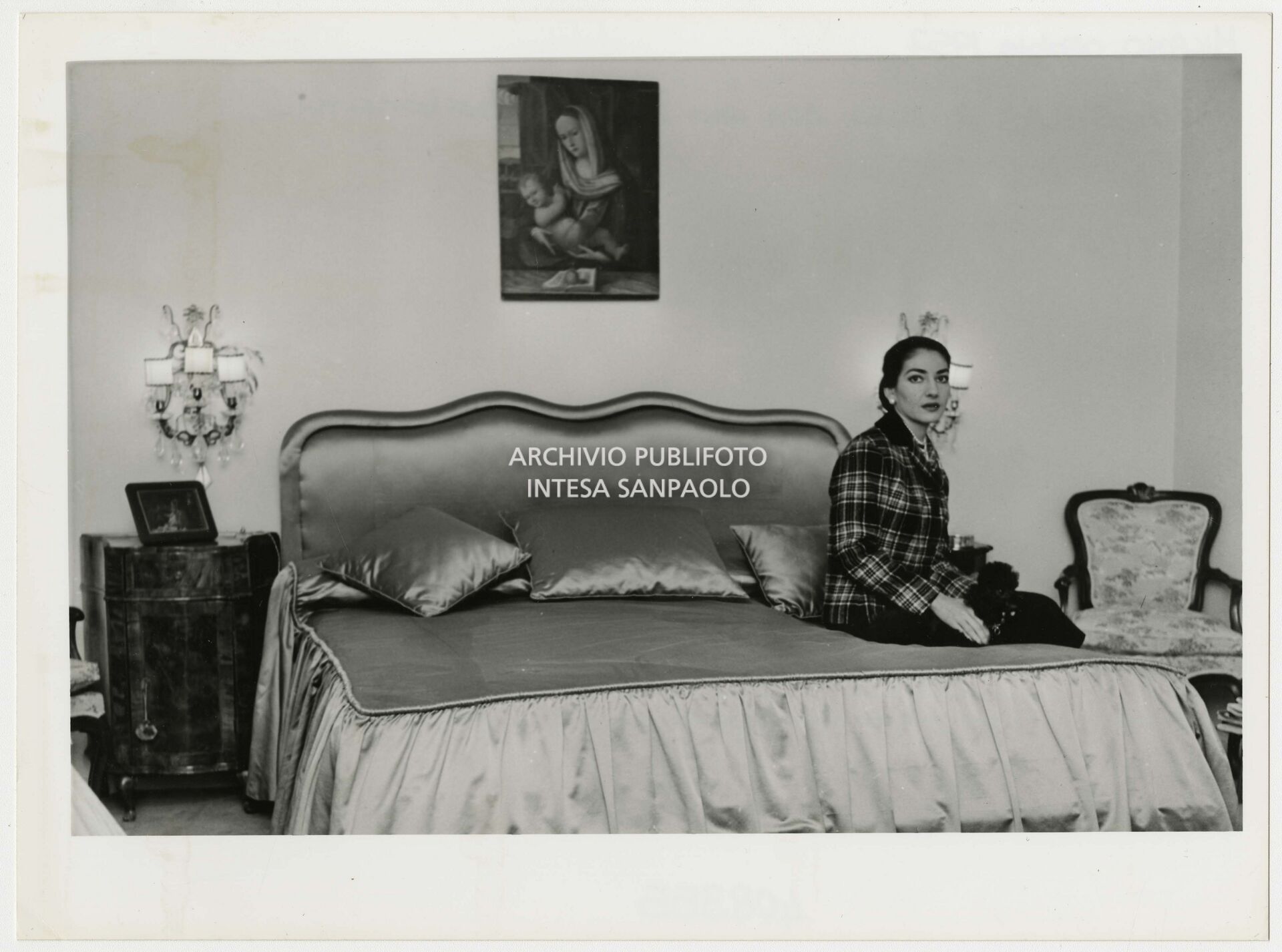Maria Callas seduta sul letto in camera con il suo barboncino; sul comodino una piccola tela, la  "Sacra Famiglia"  da lei considerato un portafortuna che portava sempre con sé