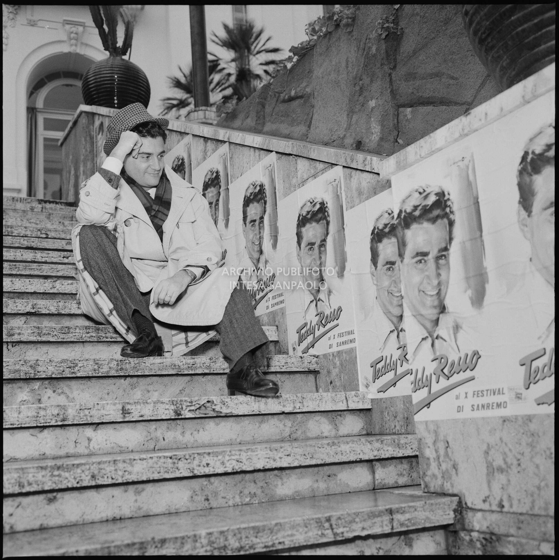 Teddy Reno posing on the Casino steps next to promotional posters featuring him during the 10th Sanremo Festival