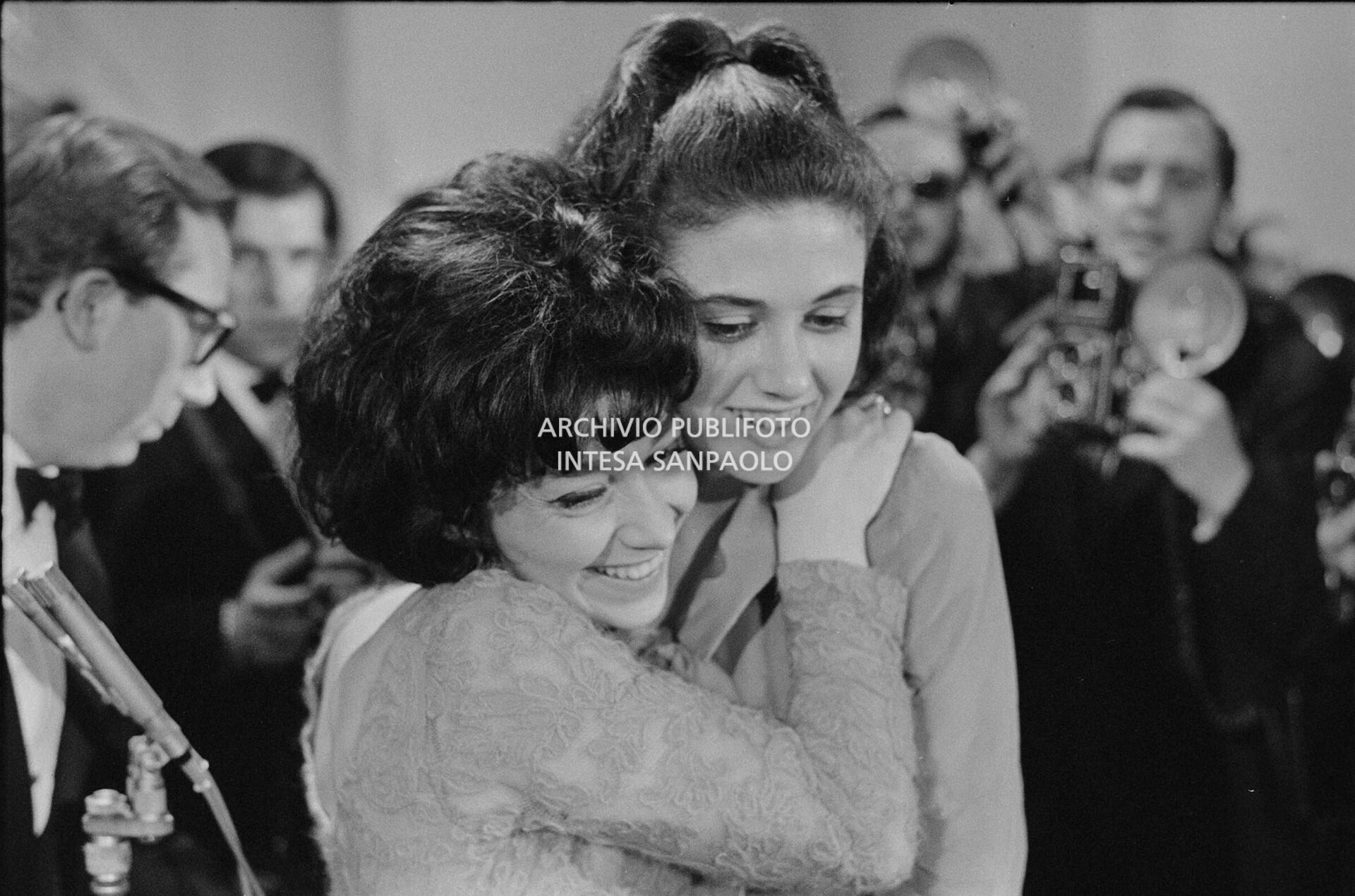 Gigliola Cinquetti e Patricia Carli, vincitrici  del XIV Festival di Sanremo, con la canzone "Non ho l'età", si abbracciano; alle loro spalle Mike Bongiorno
