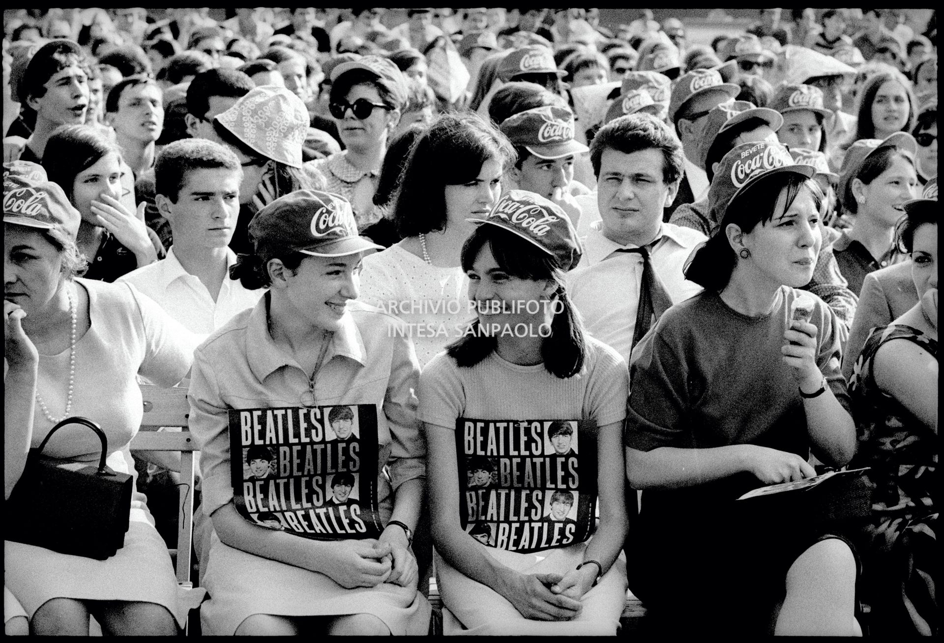 Due ragazze tra il pubblico al concerto pomeridiano dei Beatles al Vigorelli di Milano con un cartello sulla maglietta con le foto dei Beatles e il loro nome