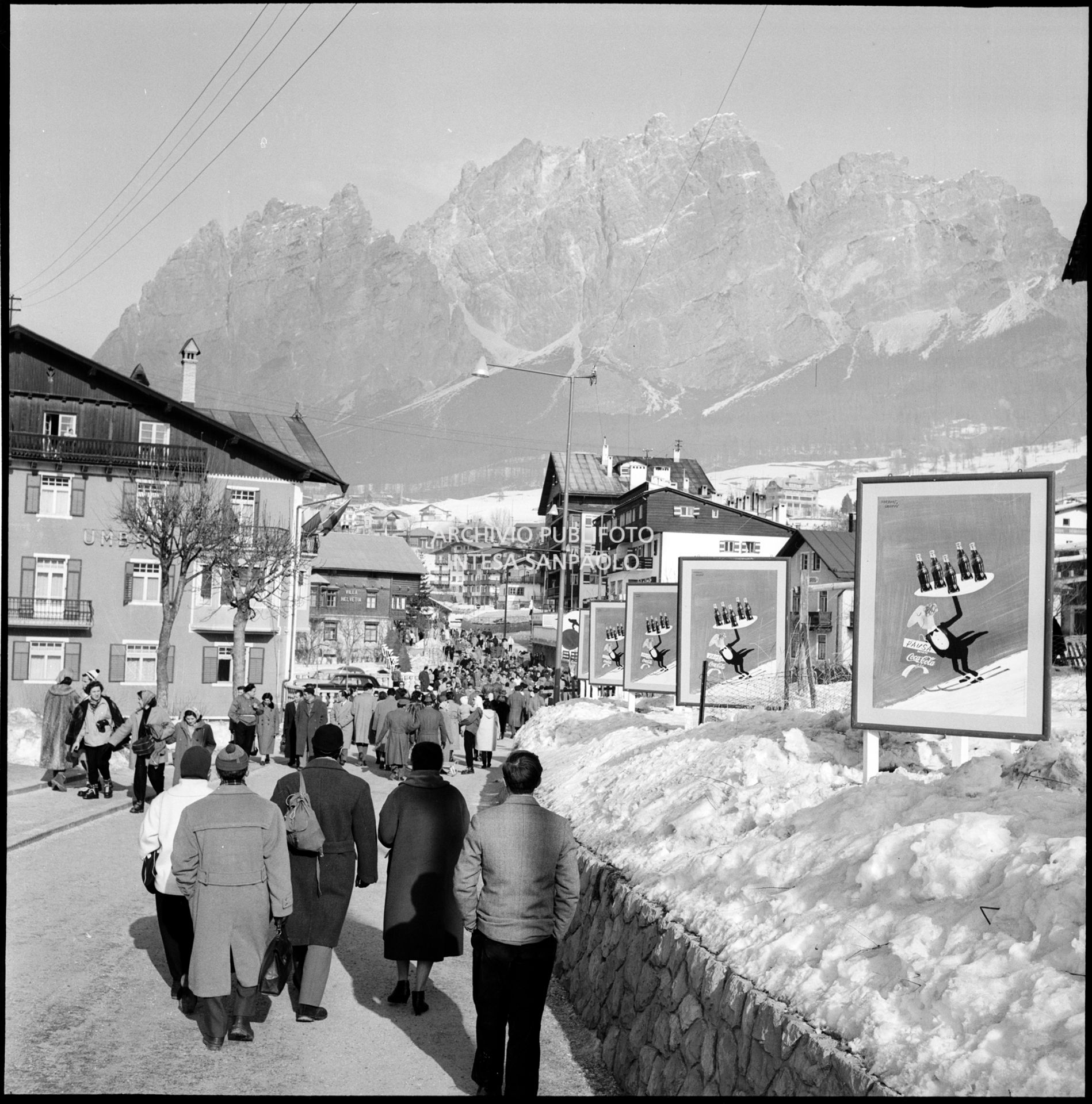 Cartelli pubblicitari Coca-Cola lungo via del Castello, affollata di passanti, a Cortina d'Ampezzo nel periodo dei VII Giochi olimpici invernali
