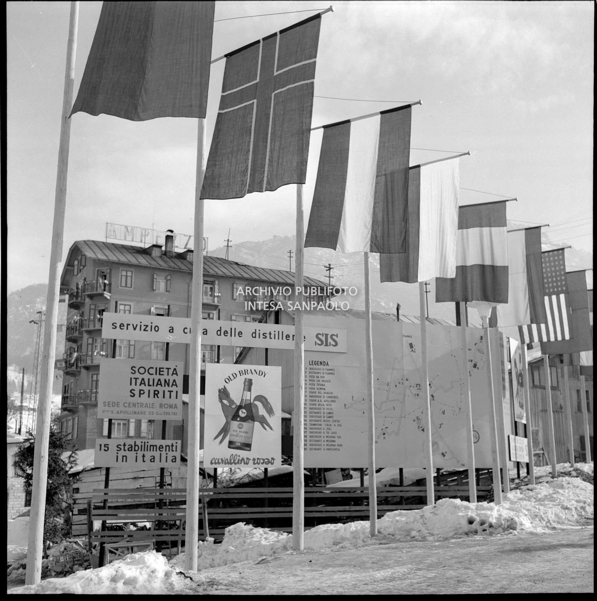 Cartelli pubblicitari della Società Italiana Spiriti, tra cui quella dell'Old Brandy Cavallino Rosso, esposti sotto le bandiere di varie nazionalità nei giorni dei VII Giochi olimpici invernali a Cortina d'Ampezzo